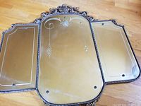 Overall view of the antique triptych-style mirror laid flat, showing three connected mirrored panels with decorative metal filigree frame and etched glass design.