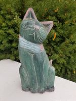 Front view of the green wooden cat statue with pink collar and facial features, showing visible cracks and textured wood grain.