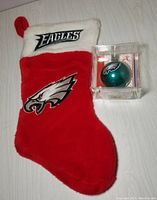 Full view of red plush Christmas stocking with white cuff and Eagles logos next to green glass Eagles ornament inside clear display box.