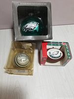 Three Philadelphia Eagles Christmas ornaments, two green round ornaments with logo and one silver round ornament with Eagles text and logo, all in original packaging.