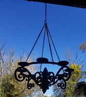 Photo of black iron candle chandelier hanging outdoors; shows decorative scrollwork and fleur-de-lis accent, suspended with chain
