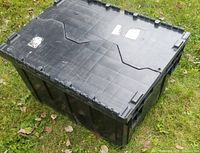 Four large black flip-top crates shown stacked outdoors on grass. Lid of top crate closed, showing wear and some stickers.