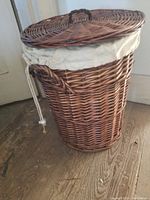 Wicker laundry hamper with lid closed, showing woven texture and fabric liner edges with drawstring handle.
