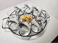 Top-down view of 8 clear glasses with black and white design placed in black iron circular carrier with handles on white surface