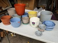 Assorted plastic planters and matching saucers of varied colors and shapes, placed on white table.