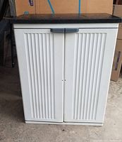 Front view of light grayish-white plastic cabinet with vertically ribbed double doors and a gray handle, topped with black stone-look surface.