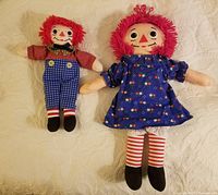 Both Raggedy Ann and Andy dolls displayed side by side on a beige blanket, showing full body and clothing details.