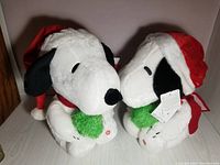 Two Snoopy plush figures side by side wearing Santa hats and red scarves holding green Christmas trees.