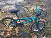Teal Mongoose Fling 100 BMX bike side view on ground with trick pegs on wheels, showing frame, seat, tires, handlebars.