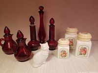 Group image showing vintage ruby red Avon cruets and bud vases, white ceramic cat-shaped bottle, and three Country Garden ceramic canisters with floral decals and cream lids.