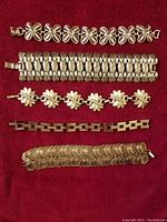 Full view of five silver-tone bracelets arranged horizontally on red fabric, showing variety of designs including chain links, florals, and coil patterns.
