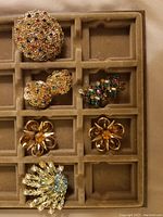 Top view of six vintage jewelry pieces on beige velvet tray, including clip-on earrings and pins with multicolored stones and gold-colored wire designs.
