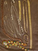 Seven pieces of vintage necklaces and bracelet sets laid out on a brown surface, including silver-colored multi-strand necklaces, gold-colored chain necklaces and bracelets, and a copper-tone necklace with enamel discs.