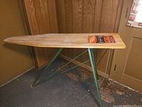 Photo showing the full wooden ironing board with green metal legs on a floor with walls and a door in the background. The top features wear and the original branding label.