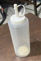 Photo of one clear plastic squeeze bottle with white twist cap and attached spout cover on a wooden surface.