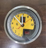 Front view of the Scuderia Ferrari RPM wall clock showing the yellow and black tachometer style dial with the Ferrari logo and digital RPM reading display.