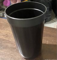 Front and angled view of a large black plastic cup on a wooden surface with a cluttered background.