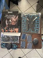 Photo showing EMS branded box with easy massage intelligent fitness equipment, two EMS muscle stimulators with blue gel pads in shape of abdominal and arm muscles, control units and loose electrode pads