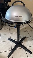 Full view of the George Foreman grill with silver dome lid on black stand, showing handle on lid and brand name text on lid.