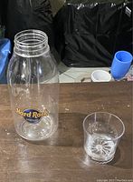 Clear plastic beverage carafe and matching glass with Hard Rock Stadium logo shown on a wooden surface.