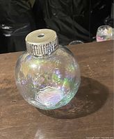 Clear spherical ornament-shaped cup with iridescent rainbow sheen and floral embossing, viewed from front on wooden surface. Silver screw-top lid is ribbed with central hole, no straw inside.