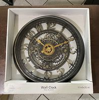 Front view of Mainstays bronze gear-style wall clock in white box showing clock face with bronze gears and numerals.