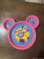 Top view of pink Minnie Mouse shaped plate with three sections showing colorful Minnie Mouse and Mickey Mouse image in the center and heart and bow decorations on ears.