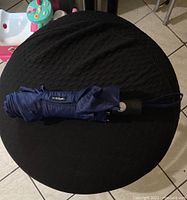 Blue Totes umbrella folded and placed on a black fabric surface, showing the arm and handle with a wrist strap.