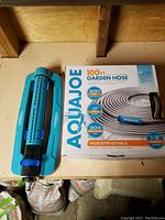 Aqua Joe 100 ft steel garden hose in box with Aqua Joe green adjustable sprinkler beside it on a wooden shelf.