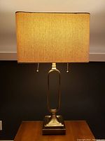 Front view of lamp showing rectangular shade and brass tone open oval body with pull chains hanging from lamp sockets.
