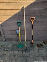 Photo showing all three tools standing against an exterior wall: Dutch hoe with green blade on the left, weed puller with wooden handle and forked metal prongs in the middle, orange-handle garden bed edger on the right.