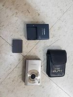 Photo showing the silver Canon digital camera front, battery charger and black carrying case arranged on floor.