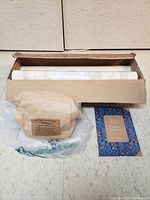 Two full wallpaper rolls and three partial rolls in a cardboard box, paper bag labeled 'Paper Hangings' wheat paste, plastic bag, and wallpaper installation book with blue floral cover.
