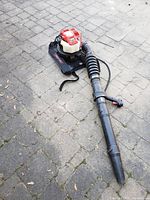 Full view of gas backpack leaf blower laid on concrete surface displaying the blower unit, flexible tubing, and harness straps.