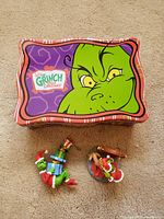 Original decorative tin box with Grinch face and logo, along with two Hallmark Grinch figurine ornaments on carpet.