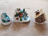 Three Hallmark Christmas ornaments arranged side by side on carpet, showing a gingerbread house marked 2002 and two winter mountain scene ornaments with icy water and skaters.