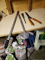 Manual hedge trimmer with long aluminum handles and black grips; green-handled spike weeder; wooden handled pruner on a wooden shelf