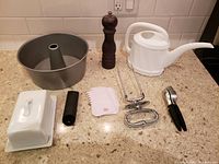 Overview of all kitchen items displayed on a kitchen counter including bundt pan, butter dish, grinder, watering can, roast holder, icing spatulas, garlic peeler, and press.