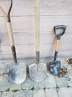 Photo showing three garden shovels with various sizes and wooden handles placed against an outdoor wall on a paved surface.