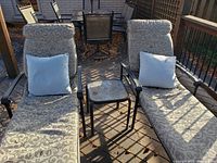 Photo of two outdoor deck lounger chairs with patterned cushions and light blue pillows, positioned on a wooden deck with a small square metal side table between them. Sunlight and shadows visible with some litter on the ground.