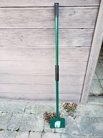 Full view of the green metal ice breaker tool with rectangular blade and rubber handle grips standing against a wooden backdrop.
