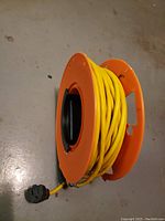 Side view of yellow extension cord on orange reel with three-prong plug visible.