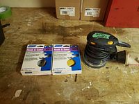 Ryobi orbital sander with two packs of Norton sanding discs on wooden surface.