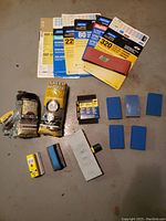 Photo showing various packages of Norton sandpaper sheets, steel wool packs, sanding blocks, sanding sponges, and a metal sanding tool arranged on the floor.