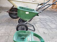 Side view of metal wheelbarrow behind green fertilizer spreader on paved surface, showing some rust on the wheelbarrow's metal frame and dirt on spreader