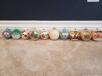 Ten assorted Christmas ball ornaments arranged in a row on beige carpet, showcasing different festive themes, colors, and designs.