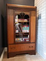 Full view of the vintage Art Deco style cabinet standing on the floor next to a brick wall, showing glass door with decorative muntins and a drawer at the bottom.