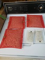 Photo showing three large red mesh bags with volcanic rock and two smaller white mesh bags arranged on a white surface with washing machine dials visible in background.