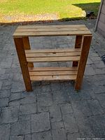 Wood shelf with three wooden plank shelves and four vertical supports placed on a paved area outdoors.
