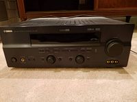 Front view of black Yamaha AV receiver showing power button, headphone jack, input selector, tuning control knobs, and volume control, with unit on carpet.